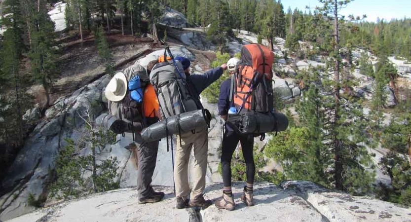three people wearing backpacking gear stand on a rock while one points into the distance on an outward bound course